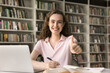 © fizkes - Cheerful beautiful college student girl showing thumb up, like hand gesture at camera, smiling, laughing, sitting at books and laptop in university library, posing for portrait