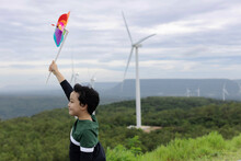 Progressive Young Asian Boy Playing With Wind Pinwheel Toy In The Wind Turbine Farm, Green Field Over The Hill. Green Energy From Renewable Electric Wind Generator. Windmill In The Countryside Concept