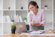 © David - Asian businesswoman working on a laptop computer at her desk in a bright modern office, doing calculating expense financial report finance making notes paper graph data document.