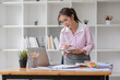 © David - Asian businesswoman working on a laptop computer at her desk in a bright modern office, doing calculating expense financial report finance making notes paper graph data document.