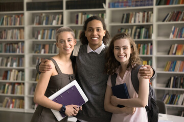 Wall Mural - Happy diverse high school friends enjoying meeting in campus library, posing with books on shelves in background, hugging, looking at camera, smiling, enjoying friendship, education