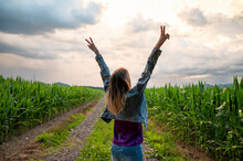 Young Woman Standing In The Middle Of Green Summer Corn Field With Her Arms Raised High In Victorious Gesture