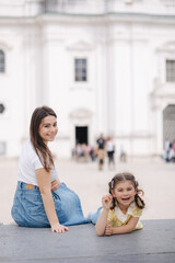 Wall Mural - Portrait of pretty young woman with her adorable daughter sitting outdoor on stone bench