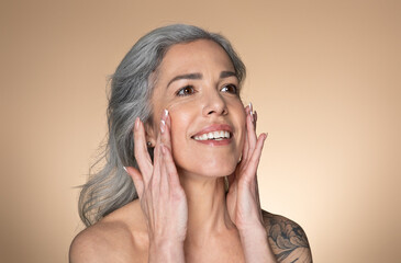 Wall Mural - Beautiful senior woman touching her smooth face skin, applying moisturizer cream or lotion on cheeks, beige background