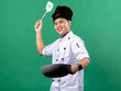 A portrait of an Indonesian Asian man wearing a chef's uniform and a chef's hat, posing with great joy, hands raised up holding a spatula and a frying pan, isolated with a green background.