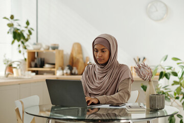 Aufkleber - Young serious Muslim businesswoman sitting by glass table in the kitchen and looking at laptop screen while scrolling through online data