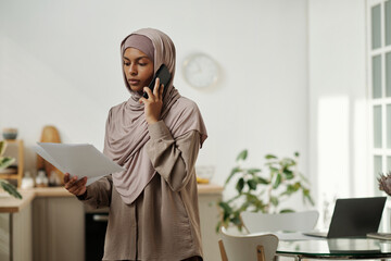 Leinwandbilder - Young female economist in hijab looking through business contract and talking to colleague or client on mobile phone in home environment