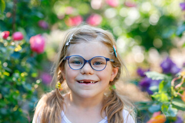 Wall Mural - Portrait of a cute preschool girl with eye glasses and big teeth gap outdoors in park. Happy funny child on sunny summer day. Kid loosing milk tooth