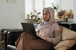 © pressmaster - Young pretty African American woman in hijab typing on laptop keyboard while sitting on couch in living room and working over new project