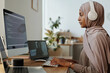 © pressmaster - Side view of young female programmer in headphones and hijab decoding online data while communicating in video chat with colleague