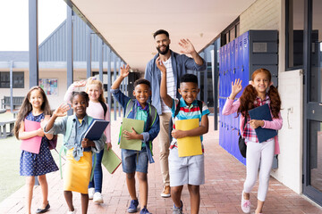 Diverse, happy male teacher and elementary schoolchildren waving in school corridor, copy space