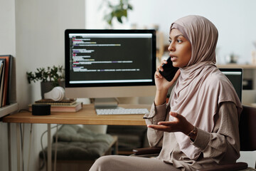 Leinwandbilder - Young confident Muslim female programmer speaking on mobile phone while explaining something to colleague against computer screen