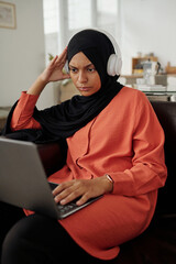Aufkleber - Young Muslim female home office worker in headphones watching online video on laptop screen while sitting on couch in front of camera
