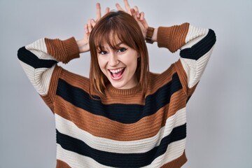 Poster - Young beautiful woman wearing striped sweater over isolated background posing funny and crazy with fingers on head as bunny ears, smiling cheerful