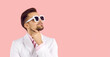 Portrait of stylish handsome young man in trendy suit and sunglasses standing on pink banner background, looking up at copy space and thinking about something with hand on chin