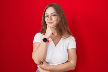 Sticker - Young caucasian woman standing over red background looking confident at the camera smiling with crossed arms and hand raised on chin. thinking positive.