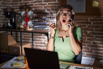 Wall Mural - Young beautiful woman working at the office at night speaking on the phone amazed and surprised looking up and pointing with fingers and raised arms.