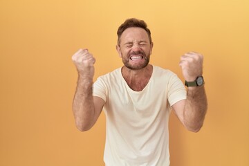 Poster - Middle age man with beard standing over yellow background very happy and excited doing winner gesture with arms raised, smiling and screaming for success. celebration concept.