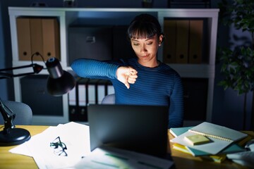 Wall Mural - Young beautiful woman working at the office at night looking unhappy and angry showing rejection and negative with thumbs down gesture. bad expression.