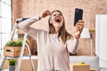 Canvas Print - Beautiful woman holding keys of new home showing on video call angry and mad screaming frustrated and furious, shouting with anger looking up.