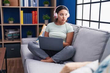 Sticker - Hispanic young woman using laptop at home wearing headphones looking to side, relax profile pose with natural face with confident smile.