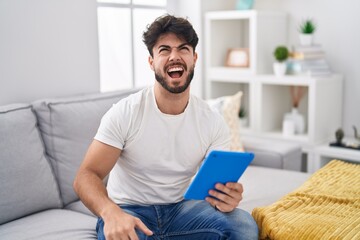 Wall Mural - Hispanic man with beard using touchpad sitting on the sofa angry and mad screaming frustrated and furious, shouting with anger. rage and aggressive concept.