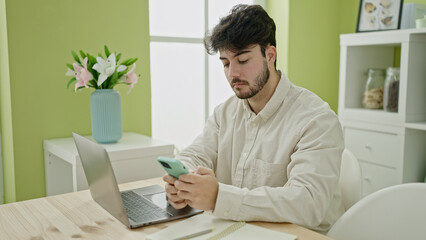 Poster - Young hispanic man using laptop and smartphone sitting on table at dinning room