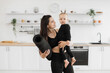 Smiling brunette woman holding barefoot infant girl and rubber mat in arms on kitchen background. Cheerful young mother and cute daughter in black sportswear preparing for yoga session at home.