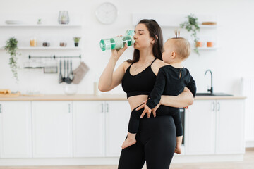 Wall Mural - Healthy young lady in yoga clothes drinking water from sports bottle while keeping baby on left side in kitchen interior. Beautiful slim mother maintaining hydration before starting training at home.