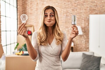 Canvas Print - Young blonde woman holding led lightbulb and incandescent bulb puffing cheeks with funny face. mouth inflated with air, catching air.