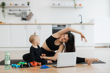 Wall Mural - Beautiful young woman and funny little girl posing on yoga mat near fitness equipment on kitchen background. Happy mom practising side bends while playful child investigating yellow resistance band.