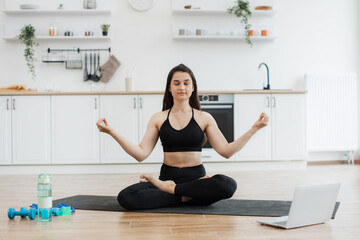 Wall Mural - Relaxed slim woman in black activewear meditating in lotus pose with hands in gyan mudra to music from laptop. Attractive lady enjoying calming tunes during yoga practice in modern apartment.