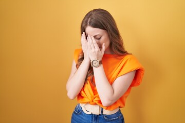 Poster - Caucasian woman standing over yellow background with sad expression covering face with hands while crying. depression concept.