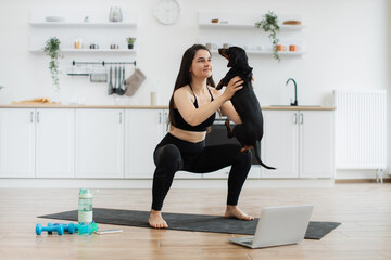 Wall Mural - Young slim female in athletic wear holding black dog in arms while crouching on yoga mat in kitchen interior. Active pet owner increasing mobility of body with perfect exercise partner at home.