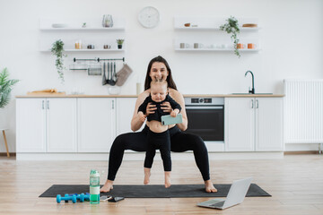 Wall Mural - Attractive caucasian adult in athletic wear practising wide squats with baby in hands in kitchen interior. Active young mother engaging cute baby daughter to physical activities indoors on sunny day.