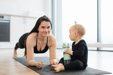 Wall Mural - Close up view of strong healthy woman in plank pose smiling at infant girl on yoga mat playing with sports bottle. Active young mom heightening baby's mood while enjoying physical training indoors.