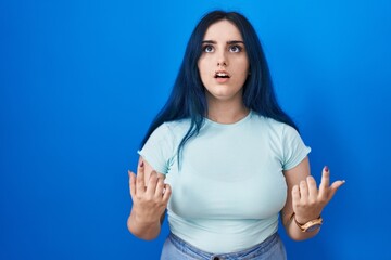 Wall Mural - Young modern girl with blue hair standing over blue background amazed and surprised looking up and pointing with fingers and raised arms.