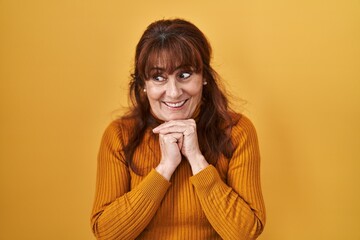 Wall Mural - Middle age hispanic woman standing over yellow background laughing nervous and excited with hands on chin looking to the side