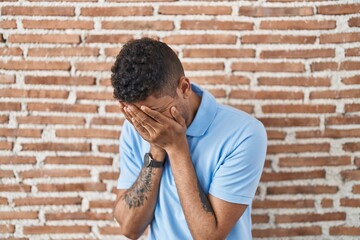 Poster - Brazilian young man standing over brick wall with sad expression covering face with hands while crying. depression concept.