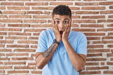 Sticker - Brazilian young man standing over brick wall afraid and shocked, surprise and amazed expression with hands on face