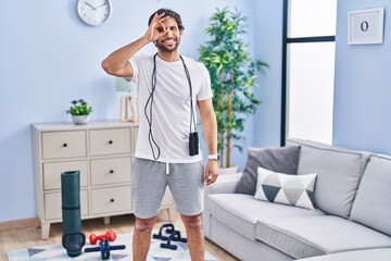 Sticker - Handsome latin man wearing sportswear at home doing ok gesture with hand smiling, eye looking through fingers with happy face.