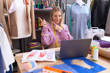 Poster - Young caucasian woman dressmaker designer using laptop smiling and looking at the camera pointing with two hands and fingers to the side.