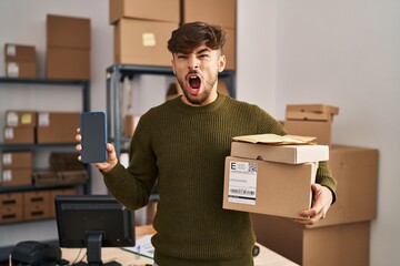 Sticker - Arab man with beard working at small business ecommerce holding delivery packages angry and mad screaming frustrated and furious, shouting with anger. rage and aggressive concept.