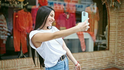 Wall Mural - Young beautiful hispanic woman smiling confident making selfie by the smartphone at clothing store