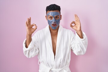 Canvas Print - Young hispanic man wearing beauty face mask and bath robe looking surprised and shocked doing ok approval symbol with fingers. crazy expression