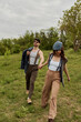 © LIGHTFIELD STUDIOS - Fashionable brunette woman in vintage outfit and newsboy cap holding hand of bearded boyfriend in sunglasses with jacket and walking on grassy field, fashionable couple surrounded by nature