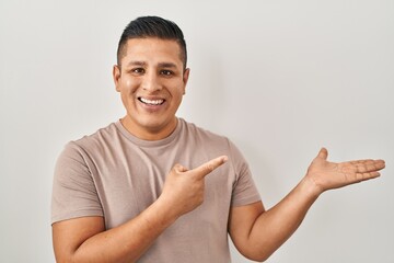 Sticker - Hispanic young man standing over white background amazed and smiling to the camera while presenting with hand and pointing with finger.