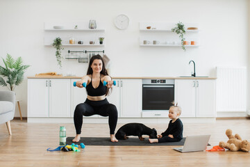 Attractive young mom practising squats with dumbbells while calm baby girl having fun with small dog on mat. Caucasian ladies in sportswear sharing daily training with canine friend in dining room.