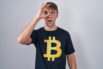 Poster - Caucasian blond man wearing bitcoin t shirt doing ok gesture shocked with surprised face, eye looking through fingers. unbelieving expression.