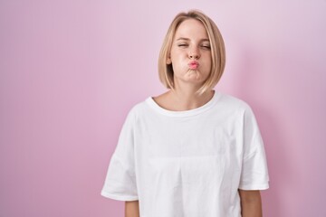 Poster - Young caucasian woman standing over pink background puffing cheeks with funny face. mouth inflated with air, crazy expression.